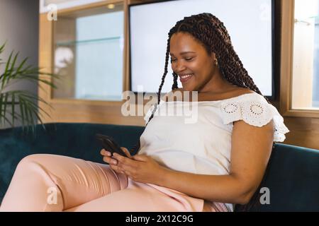 Donna birazziale con capelli intrecciati che utilizza lo smartphone in un moderno ufficio aziendale Foto Stock