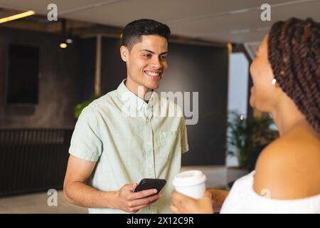 Un uomo birazziale che tiene in mano lo smartphone parla con una donna birazziale in un moderno ufficio aziendale Foto Stock