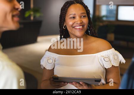 Una giovane donna afroamericana che tiene un tablet, che parla con qualcuno in un moderno ufficio d'affari Foto Stock