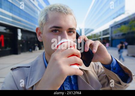 giovane biondo con capelli corti e abiti casual, al telefono, in piedi all'aperto a parlare al telefono con edifici commerciali sullo sfondo Foto Stock