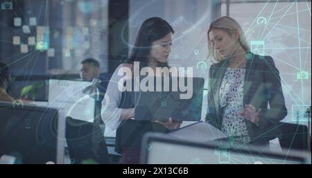 Immagine di globi di icone digitali su due donne diverse che discutono su un notebook in ufficio Foto Stock