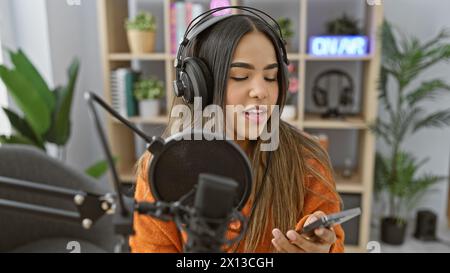 Giovane donna ispanica che canta in un moderno studio radio interno con microfono e cuffie. Foto Stock