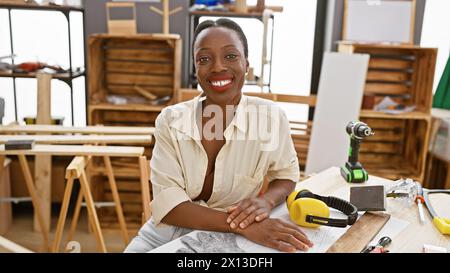 Donna afroamericana sorridente falegname seduto con fiducia al tavolo della falegnameria nel suo impegnato laboratorio al coperto Foto Stock