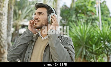 Un giovane e bello ispanico con la barba ama la musica con le cuffie all'aperto in mezzo al verde lussureggiante in un parco tranquillo. Foto Stock