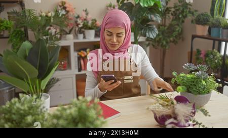 Una giovane donna che indossa un hijab e un grembiule sorride mentre usa il suo smartphone a casa, circondato da piante d'appartamento. Foto Stock