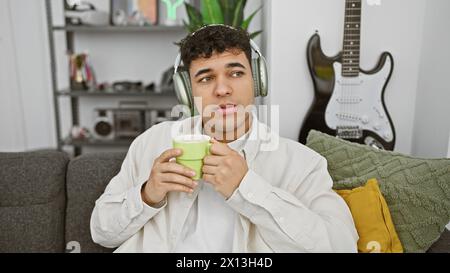 Un giovane attento si gode una bevanda calda mentre indossa le cuffie nel suo accogliente salotto con una chitarra sullo sfondo. Foto Stock