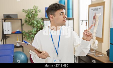 Giovane con appunti in piedi in una clinica di fisioterapia, che indica qualcosa, al chiuso. Foto Stock