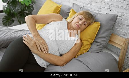 Bella donna bionda di mezza età, visibilmente turbata, sdraiata comodamente nella sua camera da letto, coinvolta nell'uso dello smartphone. ambiente interno con una mattinata accogliente Foto Stock
