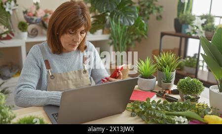 Donna matura che utilizza smartphone e laptop in un negozio di fioristi in mezzo a piante verdi in ambienti interni Foto Stock