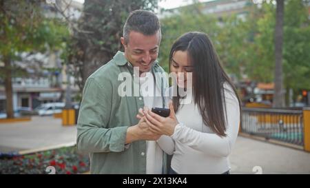 Una coppia allegra condivide un momento guardando uno smartphone insieme in un parco cittadino circondato dal verde. Foto Stock