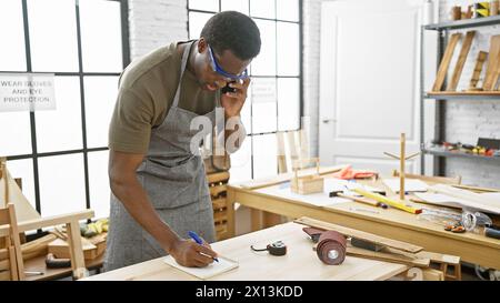 Un giovane lavora con attenzione in un'officina, indossando occhiali di sicurezza e un grembiule mentre prende appunti e fa una telefonata. Foto Stock