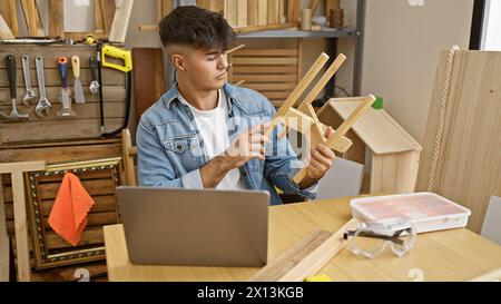 Bel giovane falegname ispanico che lavora su un oggetto di legno in un laboratorio di falegnameria, navigando abilmente in Internet sul suo portatile. Foto Stock