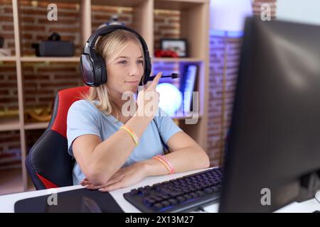 Giovane donna caucasica che gioca ai videogiochi indossando cuffie che puntano con il dito della mano a lato mostrando pubblicità, faccia seria e calma Foto Stock