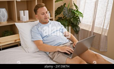 Felice e sicuro, giovane uomo caucasico si gode la sua rilassante mattinata in un'accogliente camera da letto, sorridendo piacevolmente mentre si siede su un comodo letto, esplorando l'atmosfera Foto Stock