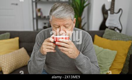 Un uomo dai capelli grigi si gode una tazza di caffè in un accogliente soggiorno con cuscini colorati e una chitarra sullo sfondo. Foto Stock