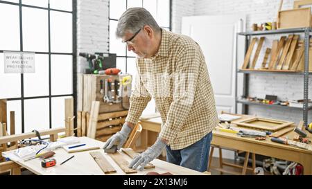 Un uomo maturo lavora diligentemente in un'officina di falegnameria ben attrezzata circondata da utensili e legno. Foto Stock