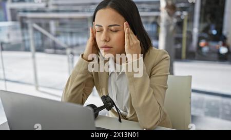 Una giovane donna stressata in abbigliamento da lavoro con gli occhi chiusi, sentendo un mal di testa durante il lavoro in un ambiente d'ufficio moderno. Foto Stock
