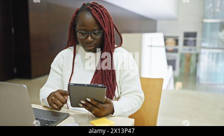 Donna afroamericana concentrata con trecce che usano un tablet in un moderno ufficio interno. Foto Stock