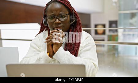 Una contemplativa donna afroamericana con trecce che indossano una cuffia in un moderno ambiente di ufficio. Foto Stock