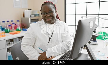 Donna afroamericana con trecce che lavora in un laboratorio, sorride con fiducia alla telecamera. Foto Stock