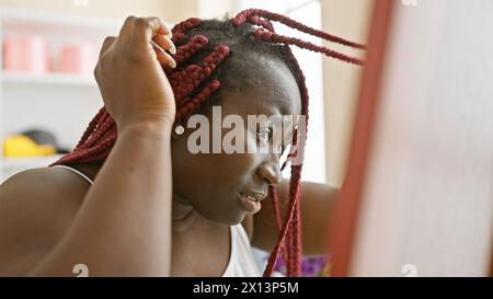 Una donna afro-americana con trecce rosse in una stanza che regola la sua acconciatura guardando lontano dalla telecamera Foto Stock