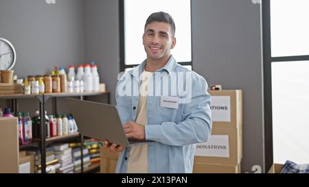 Giovane e sicuro ispanico si offre volontario con un sorriso, utilizzando un notebook in un vivace centro di beneficenza, creando unità e dando una mano. Foto Stock