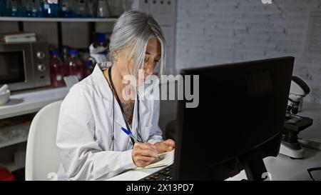 Una donna seria dai capelli grigi di mezza età, una scienziata devota, affronta il computer nel suo laboratorio, prendendo appunti diligenti in mezzo alla quiete della notte Foto Stock
