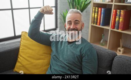 Uomo barbuto di mezza età con capelli grigi, muscoli flessibili, sorridente in un salotto moderno. Foto Stock
