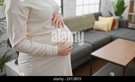 Primo piano di una donna ispanica incinta in piedi in un accogliente salotto, mano sulla pancia, simbolo della maternità. Foto Stock