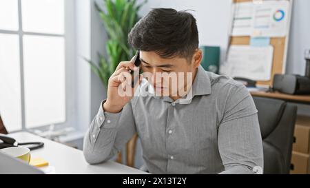 Un giovane asiatico concentrato in un ufficio parla al telefono, suggerendo affari, comunicazioni o servizio clienti. Foto Stock