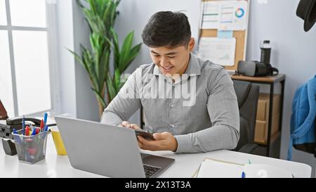 Un giovane asiatico sorride mentre utilizza uno smartphone alla sua moderna scrivania, ritraendo un ambiente di lavoro professionale. Foto Stock