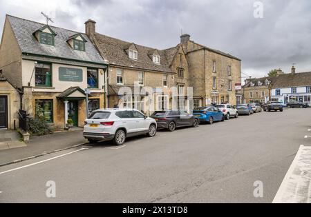 Un'affascinante Stow sulla Wold Street fiancheggiata da negozi di pietra del Cotswold e auto parcheggiate, catturando l'essenza della vita dei villaggi inglesi. Foto Stock