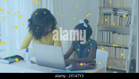 Immagine di macchie gialle che fluttuano sopra la madre afroamericana che aiuta la figlia a fare i compiti a casa Foto Stock