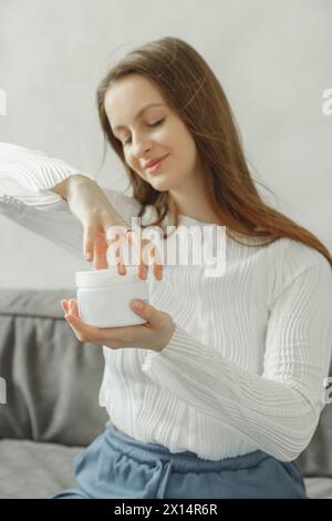 Barattolo apribile donna sorridente con crema idratante, maglione bianco, trattamenti di bellezza a casa Foto Stock