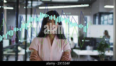 Immagine di più grafici con numeri che cambiano su una donna birazziale sorridente con le braccia incrociate Foto Stock