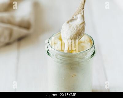 Pronto per preparare il pane di pasta di grano soursough antipasto gocciolando da cucchiaio di legno. Vaso in vetro con antipasto di pasta acida idratazione al 100% su fondo di legno bianco. Spazio di copia. Concetto di panificazione fatto in casa Foto Stock