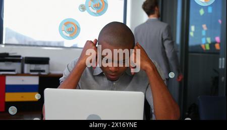 Immagine di più icone grafiche su un uomo afro-americano stanco che guarda il laptop in ufficio Foto Stock