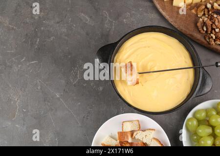 Pentola di fonduta con gustoso formaggio fuso, forchetta e diversi spuntini su un tavolo grigio, piatto. Spazio per il testo Foto Stock