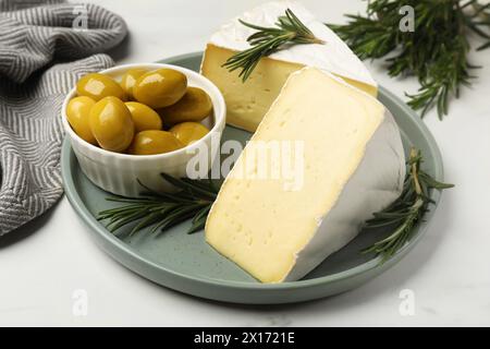 Piatto con pezzi di gustoso formaggio camembert, olive e rosmarino su tavola bianca, primo piano Foto Stock