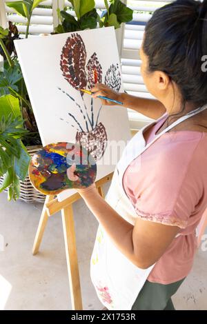 Donna birazziale matura che dipinge su tela a casa, indossando grembiule Foto Stock