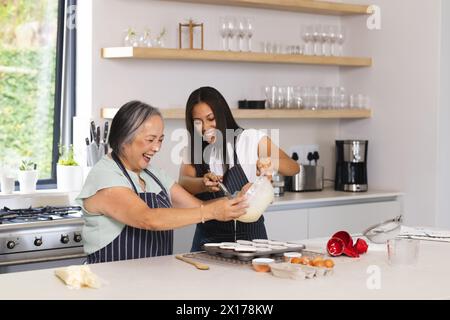 Nonna asiatica e nipote adolescente birazziale stanno cucinando insieme a casa. La nonna ha i capelli grigi e sorride, la nipote ha una lunga h nera Foto Stock