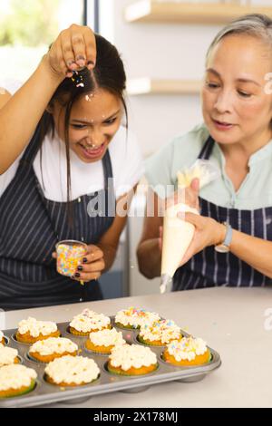 La nonna asiatica e la nipote adolescente birazziale stanno decorando i cupcake a casa Foto Stock