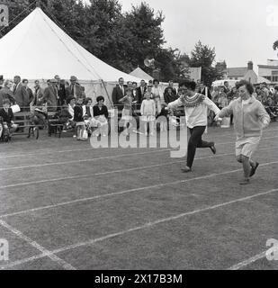 1964, storico, una corsa di uova e cucchiai per adulti, due donne all'aperto su una pista in erba che gareggiano in una divertente corsa in una giornata di attività di servizi combinati in cui devono bilanciare un uovo o un oggetto a forma di smiliar su un cucchiaio, mentre corrono con esso fino al traguardo. Foto Stock