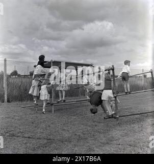 1964, storico, in un campo fuori da una tenuta abitativa, bambini piccoli che giocano su una cornice di arrampicata, due dei quali appesi a testa in giù sui pali d'acciaio. I telai da arrampicata sono una buona forma di esercizio per i giovani, migliorando la forza, l'equilibrio e il coordinamento e le prove dimostrano che i bambini che hanno giocato su di loro sono più agili e sicuri di sé. Foto Stock