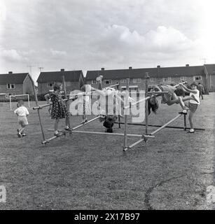 1964, storico, in un campo fuori da una tenuta abitativa, bambini piccoli che giocano su una cornice di arrampicata, due ragazze appese a testa in giù sui pali d'acciaio. I telai da arrampicata sono una buona forma di esercizio per i giovani, migliorando la forza, l'equilibrio e il coordinamento e le prove dimostrano che i bambini che hanno giocato su di loro sono più agili e sicuri di sé. Foto Stock