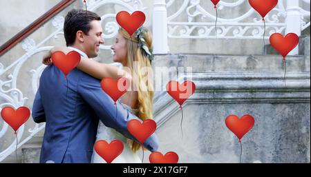 Immagine di più palloncini rossi a forma di cuore che fluttuano sopra una coppia appena sposata che balla e sorride Foto Stock