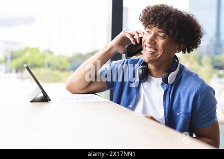 Giovane birazziale che parla al telefono, indossa le cuffie intorno al collo in un moderno ufficio aziendale Foto Stock