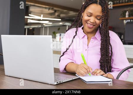 Una giovane donna birazziale che indossa una camicia rosa scrive su un notebook in un moderno ufficio aziendale Foto Stock