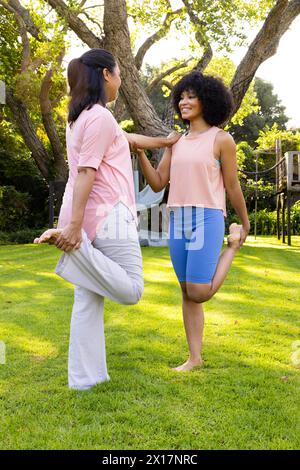 Madre e figlia birazziali che si tengono per mano, in equilibrio su un piede nel giardino di casa Foto Stock