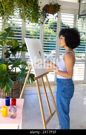 Giovane donna birazziale che dipinge su tela a casa, circondata da piante Foto Stock